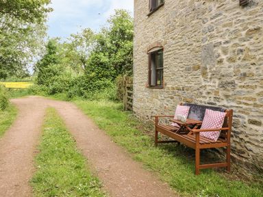 A wooden bench with cushions and a small table outside a stone house beside a dirt path and greenery at Cottage on the Common in Westbury-on-Severn