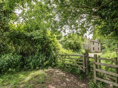 A wooden gate opening to a dirt and grassy area with dense green foliage and a stone house in the background at Cottage on the Common in Westbury-on-Severn