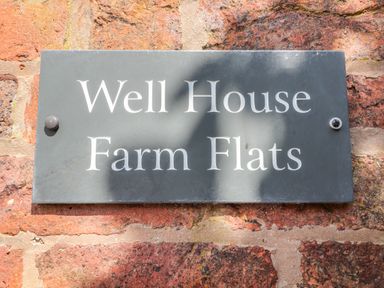 A black sign with white text reading Well House Farm Flats mounted on a red brick wall at Well House Farm Flat 1 in Bretton