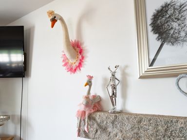 A wall with swan decorations and a silver statue on a stone mantel a dandelion framed picture and a tv nearby at Be Our Guest Cottage in Chacewater near Truro