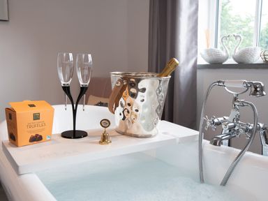 A bathtub filled with water and bubbles with a tray holding two champagne glasses a champagne bottle in an ice bucket chocolates and a small bell at Be Our Guest Cottage in Chacewater near Truro