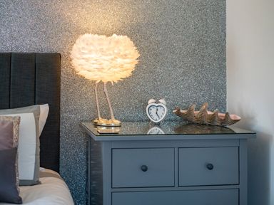 A bedside table with a feather lamp clock and decorative shell next to a bed at Be Our Guest Cottage in Chacewater near Truro