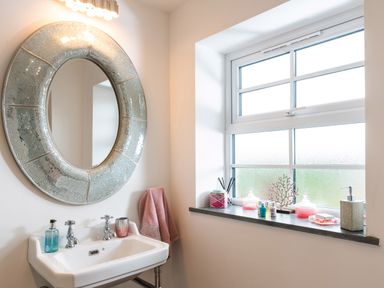 A bathroom with a sink a round mirror and a window with toiletries and decorations at Be Our Guest Cottage in Chacewater near Truro