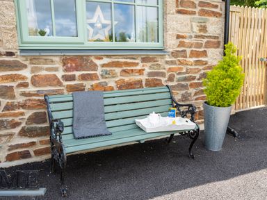 A green bench with a gray blanket and a tray with tea set outside a stone house with a green window and a potted plant at Be Our Guest Cottage in Chacewater near Truro