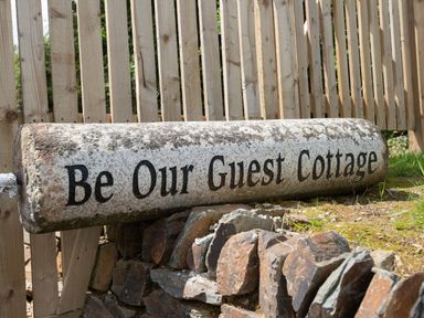 A stone sign with the text Be Our Guest Cottage near a wooden fence and rocks at Be Our Guest Cottage in Chacewater near Truro