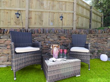 An outdoor seating area with two wicker chairs a wicker table with glasses and a pitcher on artificial grass at Be Our Guest Cottage in Chacewater near Truro