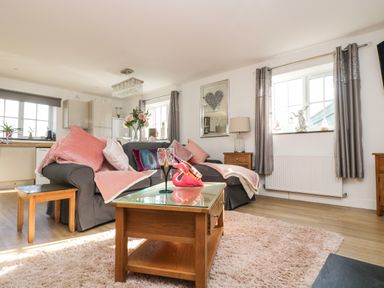 A living room with a gray sofa and pink cushions and throws a wooden coffee table with glass top a dining table with flowers and kitchen area in the background at Be Our Guest Cottage in Chacewater near Truro
