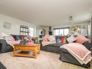 A living room with gray sofas pink pillows and throws a wooden coffee table and an open kitchen at Be Our Guest Cottage in Chacewater near Truro
