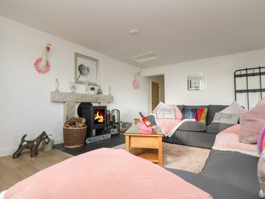 A living room with grey sofas pink cushions a wooden coffee table and a wood burning stove at Be Our Guest Cottage in Chacewater near Truro