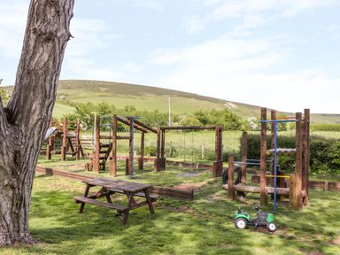 An outdoor playground with wooden climbing frames swings a picnic table and a toy car in a grassy area at Duck Cottage in Sutton Poyntz