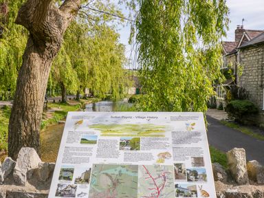 A village history information board next to a river with trees and stone houses at Duck Cottage in Sutton Poyntz