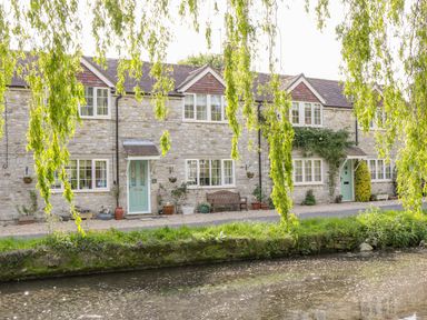 Stone cottages with pale green doors and windows by a small waterway with overhanging tree branches at Duck Cottage in Sutton Poyntz