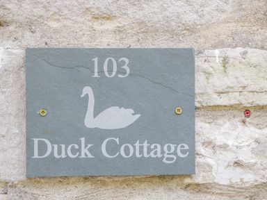 A stone wall with a gray house number plaque showing 103 and a swan above the text Duck Cottage in Sutton Poyntz