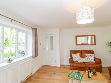 A living room with a brown leather sofa pillows a coffee table with a Scrabble board wine and snack a window with curtains and a door at Duck Cottage Sutton Poyntz