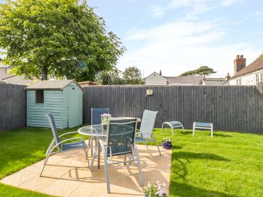 A backyard with a small light blue shed a patio with a glass table and four chairs and green grass at Duck Cottage in Sutton Poyntz