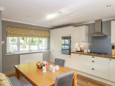 A kitchen with wooden dining table chairs white cabinets and a window with yellow roman blind at Duck Cottage in Sutton Poyntz