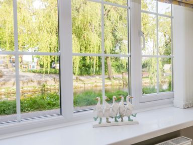 A window with white frames overlooking trees and a river with a decoration of four white ducks on the windowsill at Duck Cottage in Sutton Poyntz