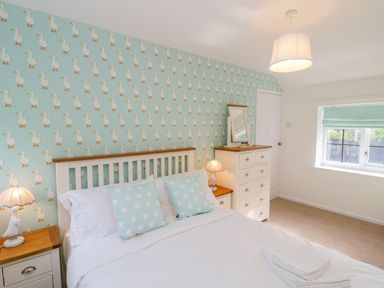 A bedroom with a white bed featuring polka dot pillows a nightstand with a lamp a chest of drawers with a mirror and duck patterned wallpaper at Duck Cottage in Sutton Poyntz