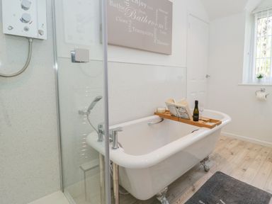 A bathroom with a clawfoot bathtub holding a wooden tray with a book a candle a bottle and a glass near a walk-in shower and a window at Duck Cottage in Sutton Poyntz