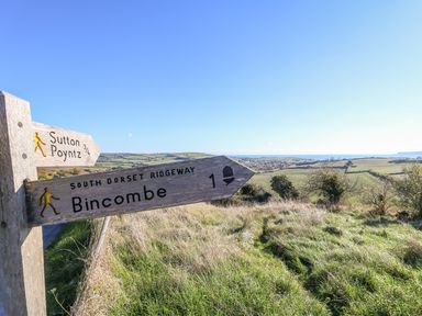 A wooden signpost on a grassy hill with directions to Sutton Poyntz and Bincombe with fields and a village in the background at Duck Cottage in Sutton Poyntz