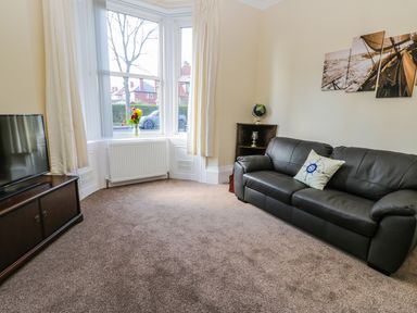 A living room with a black leather sofa a wooden corner shelf with a globe and lamp a television on a wooden cabinet and a vase with flowers on a window ledge at Sailors Lodge in Filey