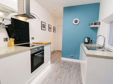 A kitchen with an oven stove and cutting board on left side a sink kettle and containers on right side a blue wall with a clock and three pictures on white wall at Sailors Lodge in Filey