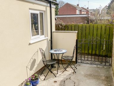 A small outdoor patio with a round mosaic table and two metal chairs next to a window and a gate with a wooden fence in the background at Sailors Lodge in Filey