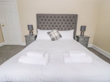 A bedroom with a bed with gray headboard white bedding two bedside tables with lamps and folded towels at Coquet View Apartment in Warkworth