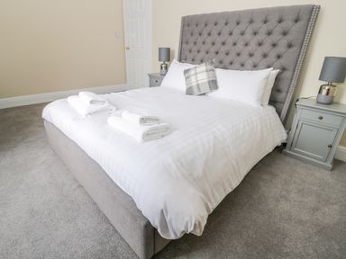 A bedroom with a gray upholstered bed with white linens and folded towels on top with bedside tables and lamps at Coquet View Apartment in Warkworth