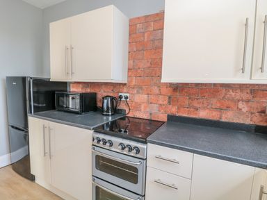 A kitchen with white cabinets a black microwave a black kettle a stove and an exposed brick backsplash at Coquet View Apartment in Warkworth