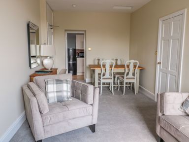 A living room with beige armchairs a wooden dining table with white chairs and a small side table with a lamp at Coquet View Apartment in Warkworth