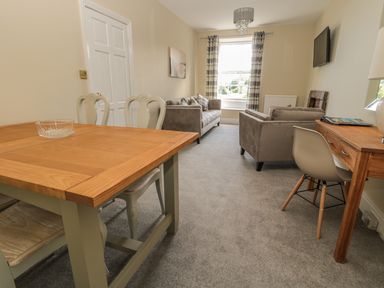 A living room with a wooden table and chairs two sofas a desk with a chair and a wall-mounted TV at Coquet View Apartment in Warkworth