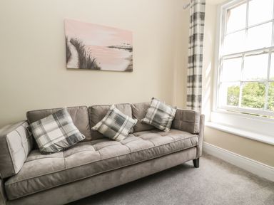 A room with a gray sofa with plaid cushions a window with plaid curtains and a landscape painting on the wall at Coquet View Apartment in Warkworth