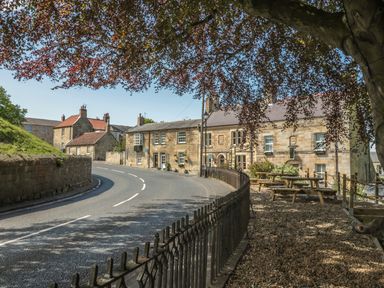 A street with stone buildings a curved road picnic tables and a tree in a village
