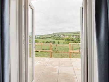 A view through open double doors onto a tiled patio and a fenced grassy area with fields and hills in the distance at Village View in Burton Bradstock
