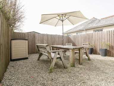 A fenced outdoor area with gravel ground a wooden table wooden chairs a bench a large umbrella and plant pots at Lily Cottage in Camelford