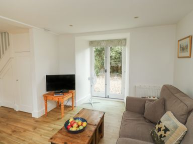 A living room with a brown sofa a wooden coffee table with a bowl of fruit a small wooden table with a television and a standing fan near glass doors at Bantam Cottage in Little Woodford near Ottery St Mary