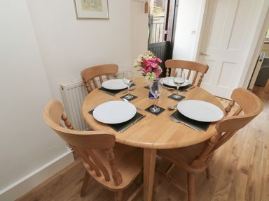 A dining area with a round wooden table set for four with plates glasses cutlery and a flower vase at Bantam Cottage in Little Woodford near Ottery St Mary