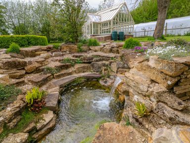 A garden pond with a small waterfall surrounded by stone walls and plants with a greenhouse in the background at Tickmorend Farm in Nailsworth