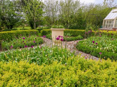 A garden with flower beds stone pathways a large planter and a greenhouse at Tickmorend Farm in Nailsworth