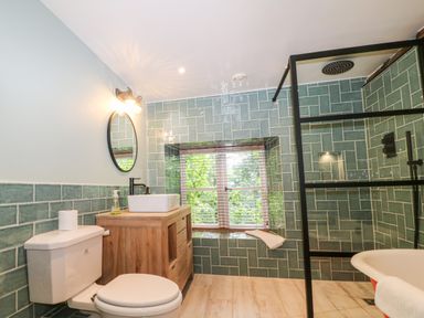 A bathroom with a toilet, wooden vanity with sink, round mirror, window with blinds, and walk-in shower at Tickmorend Farm in Nailsworth