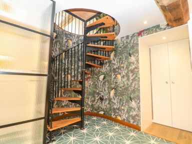 A spiral staircase with wooden steps and black metal railing in a room with patterned wallpaper and tiled floor at Tickmorend Farm in Nailsworth