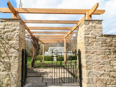 A stone wall entrance with a black iron gate and wooden pergola leading to a garden with a greenhouse at Tickmorend Farm in Nailsworth