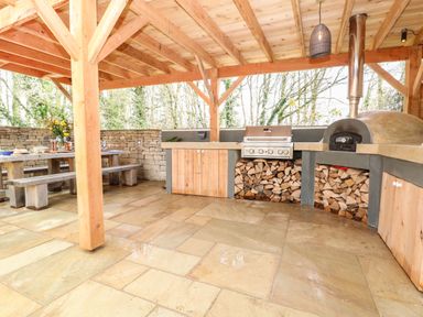 An outdoor covered kitchen area with wooden beams a pizza oven a grill firewood storage and a wooden picnic table at Tickmorend Farm in Nailsworth