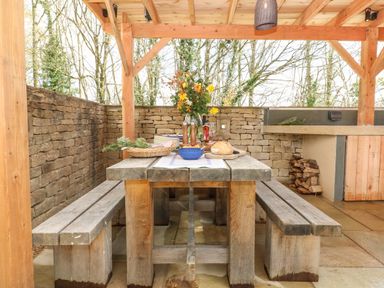 An outdoor dining area with a wooden table and benches under a wooden pergola at Tickmorend Farm in Nailsworth