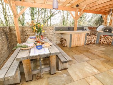 An outdoor dining area with a wooden table and benches set with food and flowers next to a stone wall and a wood-fired oven at Tickmorend Farm in Nailsworth