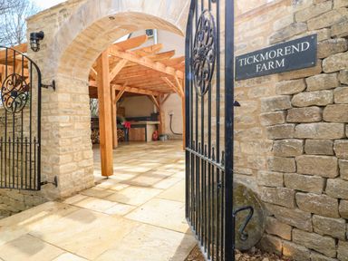 Stone archway with open wrought iron gate leading to a covered outdoor area with wooden beams and stone floor at Tickmorend Farm Nailsworth