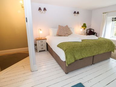 A bedroom with a bed covered by a green blanket and two decorative pillows two bedside tables with lamps a chair by the window and a doorway leading to a carpeted area at Tickmorend Farm in Nailsworth