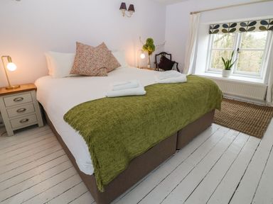 A bedroom with a large bed with green blanket and floral pillows a bedside table with lamp and a window with a plant at Tickmorend Farm in Nailsworth