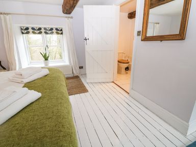 A bedroom with a green bedcover towels on the bed a window with white curtains a door leading to a bathroom with a toilet at Tickmorend Farm Nailsworth